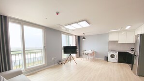 A spacious living area with a large window, a flat-screen TV on a tripod, a dining table with chairs, a washing machine, and a kitchen area.
