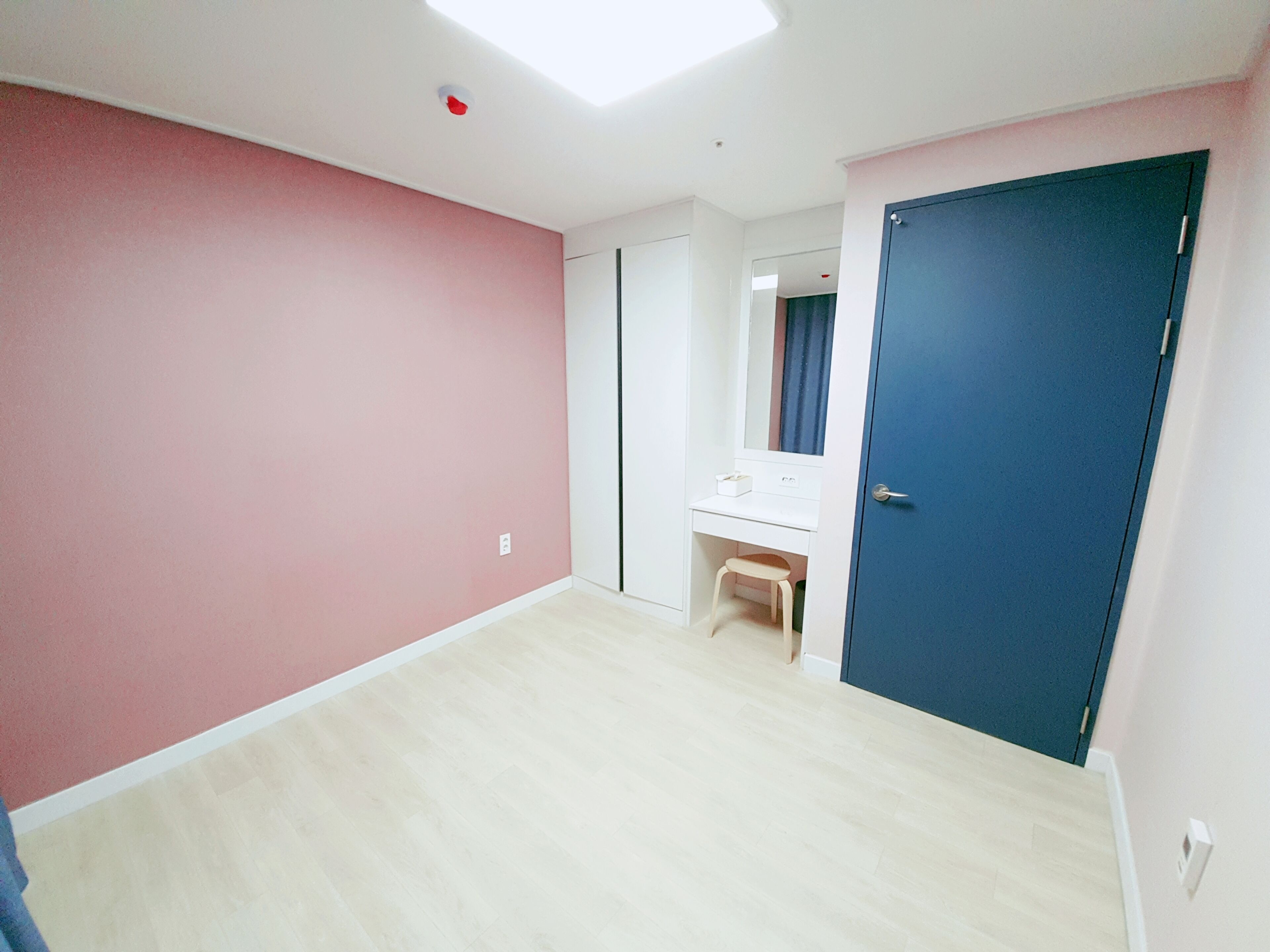 A room with pink walls, a wooden floor, a white desk with a mirror, and a blue door.