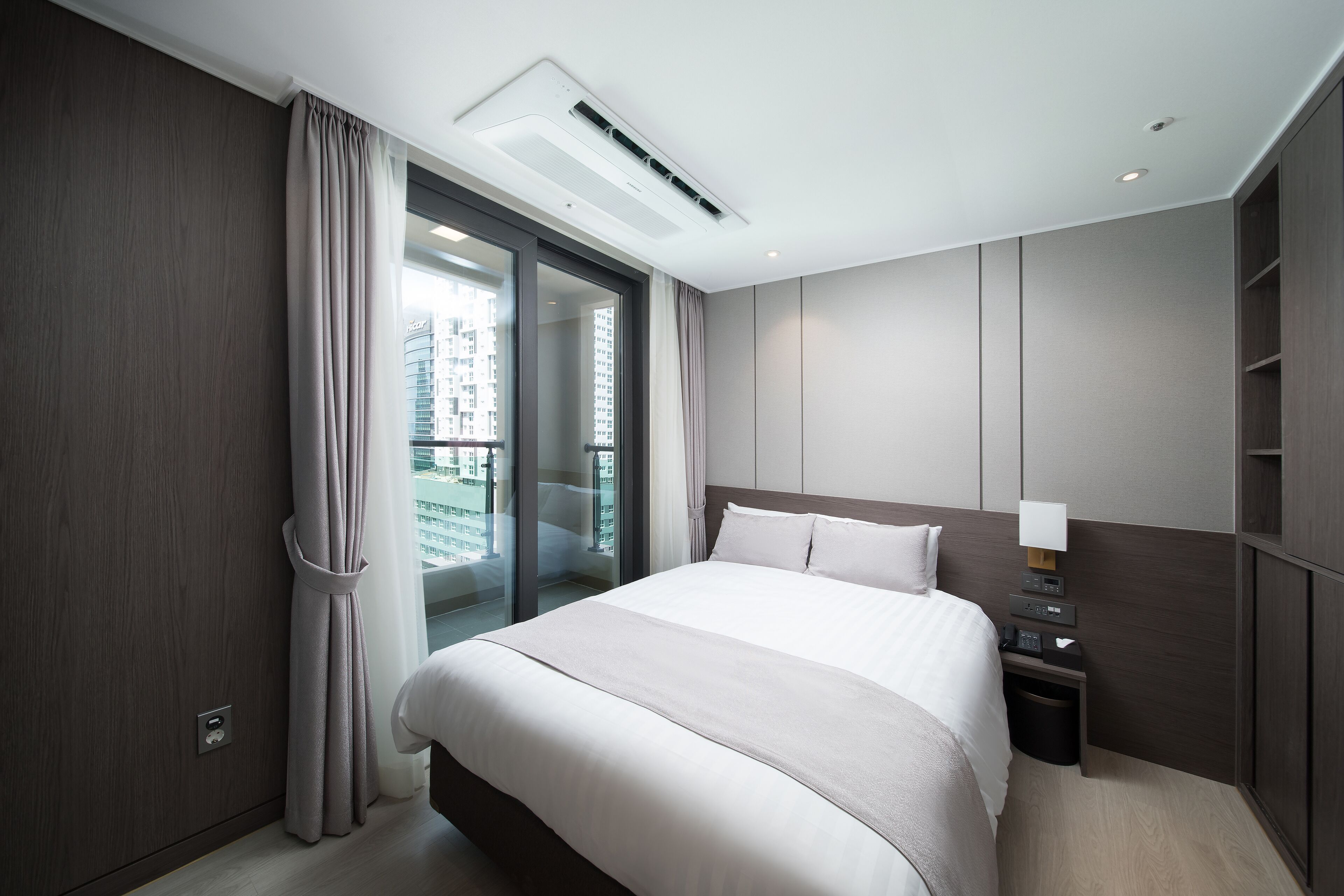 Una habitación de hotel moderna con una cama grande, una mesita de noche con lámpara, un cubo pequeño de basura y vistas a un paisaje urbano a través de un ventanal corredero.