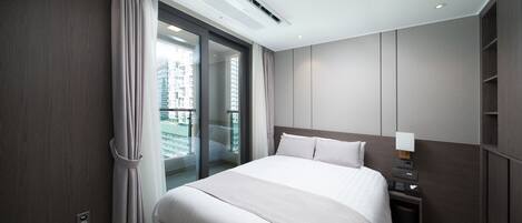 A modern hotel room with a large bed, a nightstand with a lamp, a small trash bin, and a view of a cityscape through a sliding glass door.