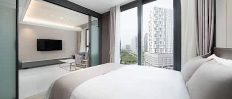 A modern hotel room with a large bed, a TV mounted on the wall, and a view of the cityscape through large windows.