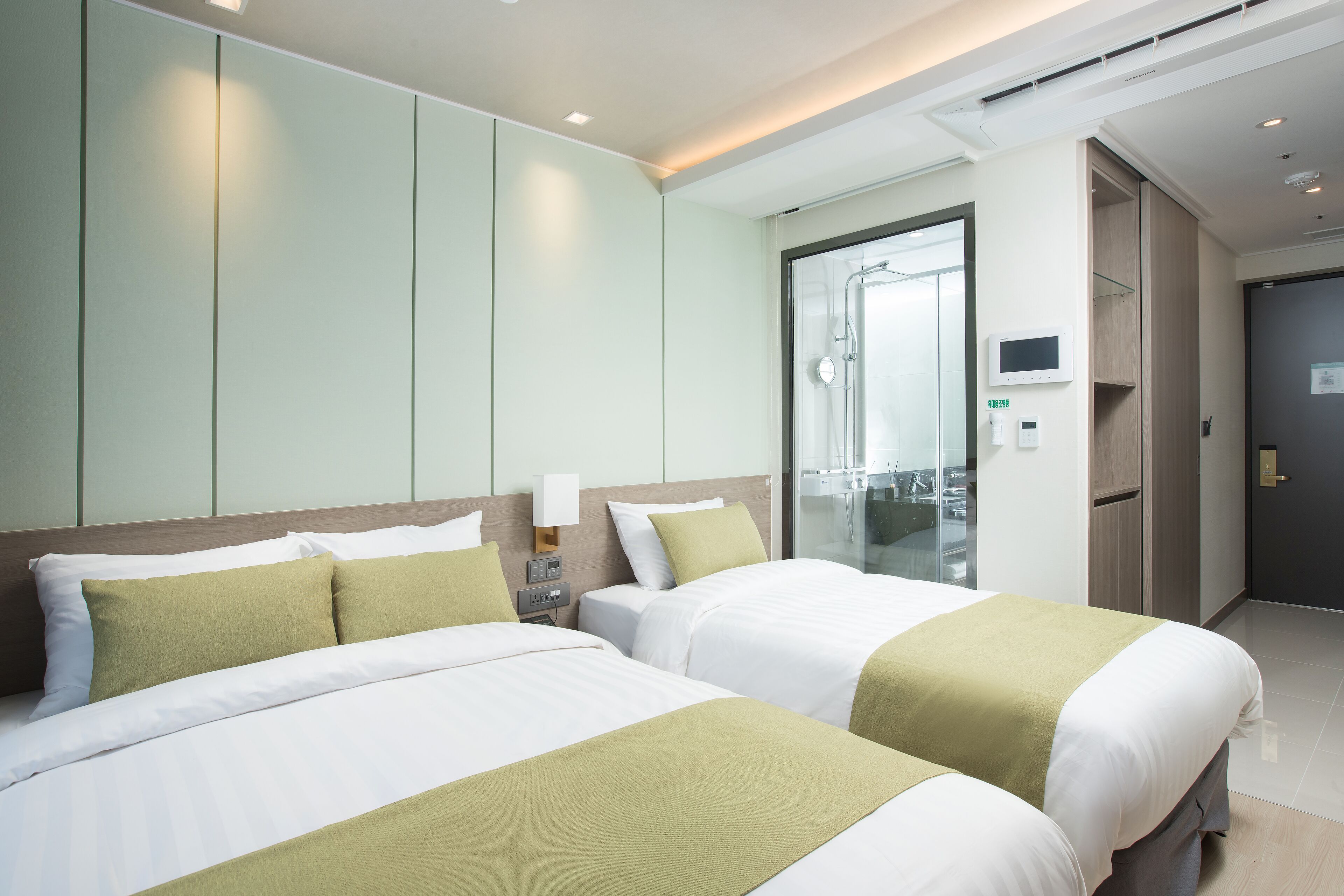 A hotel room with two beds, a bedside table, a TV, and a bathroom visible through a glass door.