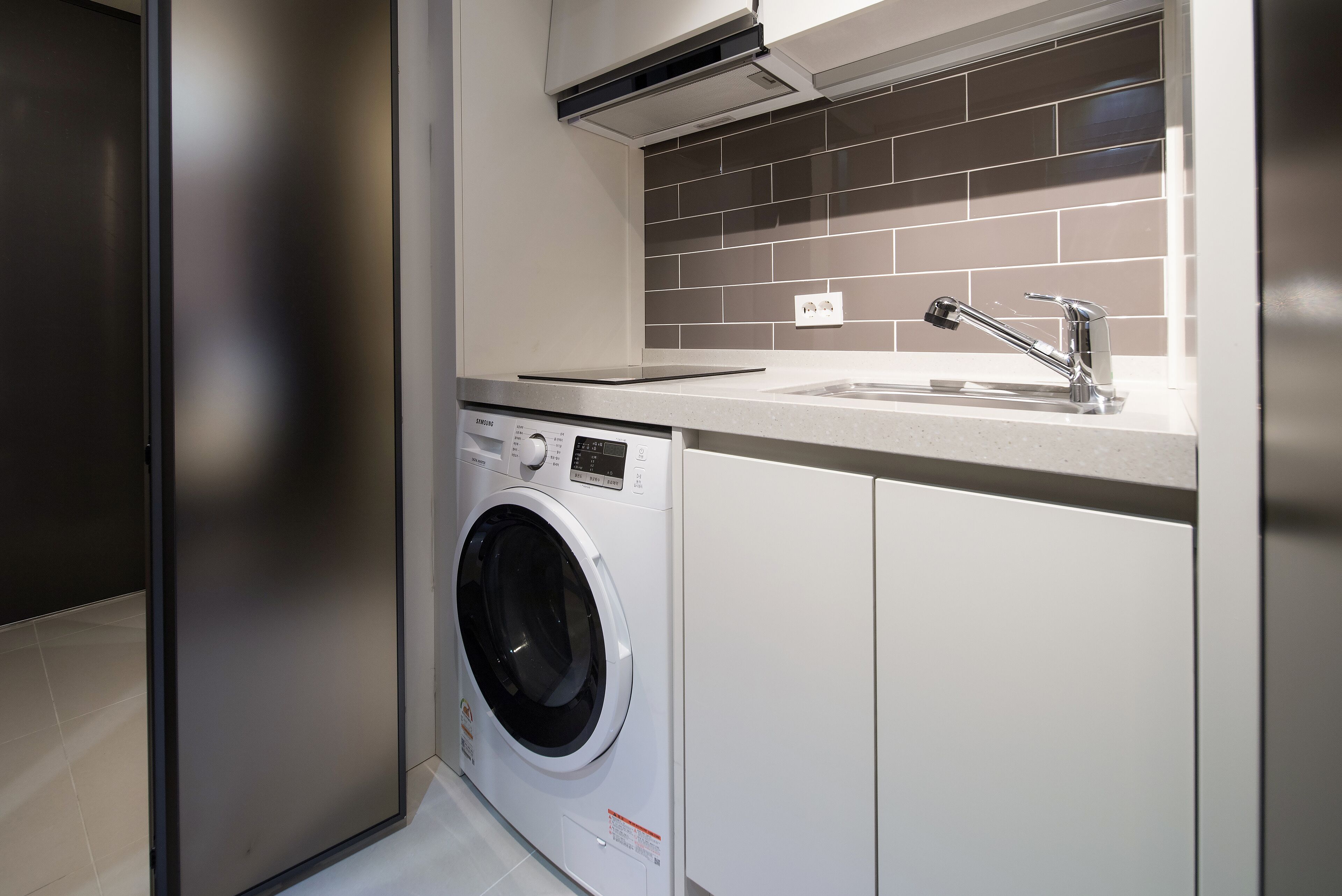 A modern laundry room with a front-loading washing machine, a sink, and a tiled backsplash.