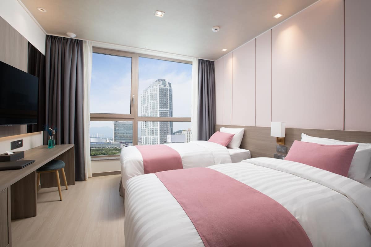 A hotel room with two beds, a desk, a TV, and a large window with a city view.