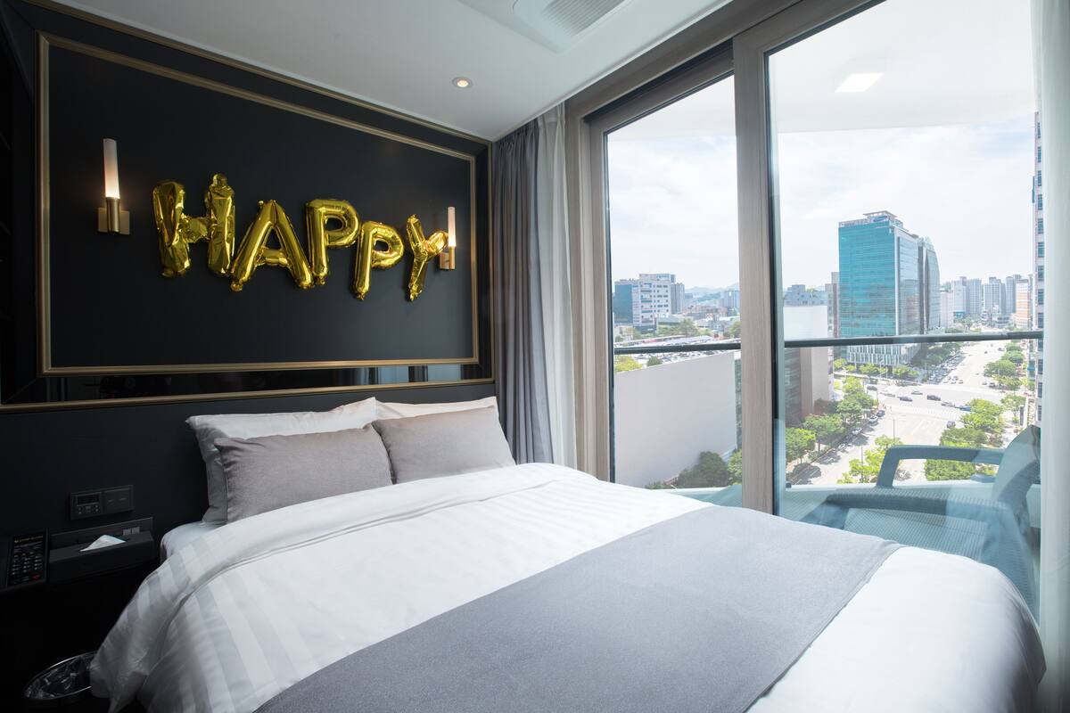 A modern hotel room with a large bed, a city view, and a 'HAPPY' balloon decoration on the wall.