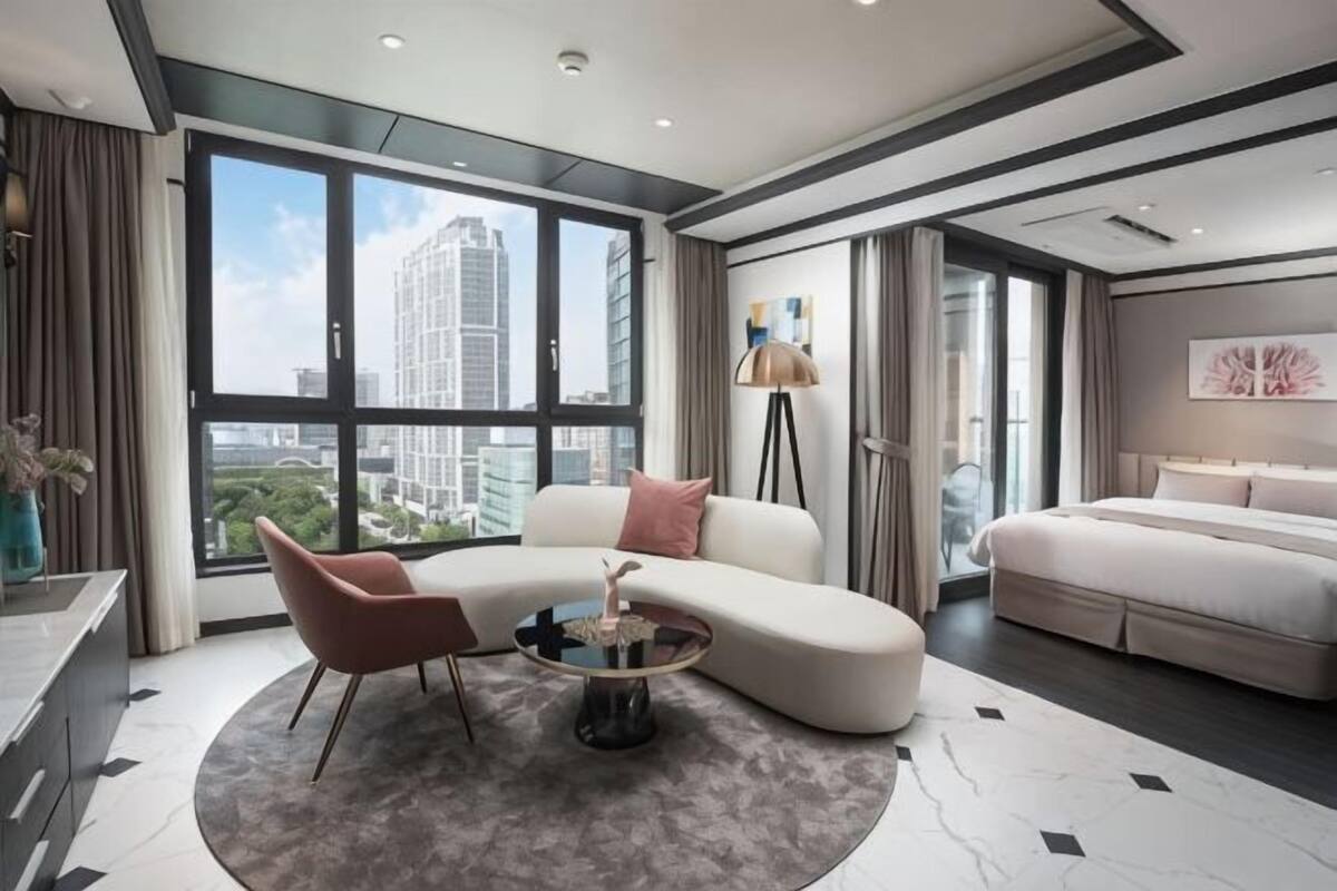 A modern hotel room with a curved sofa, a round coffee table, a bed, and a large window offering a city view.