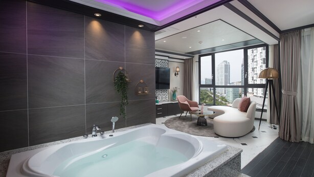 Un baño moderno con una bañera grande, paredes de azulejos oscuros y vistas de la ciudad a través de amplias ventanas.