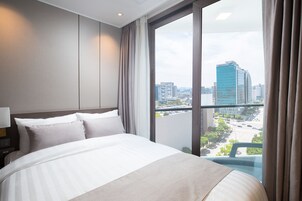 Habitación de hotel con una cama grande, una mesita de noche y vistas a un paisaje urbano a través de un ventanal amplio.
