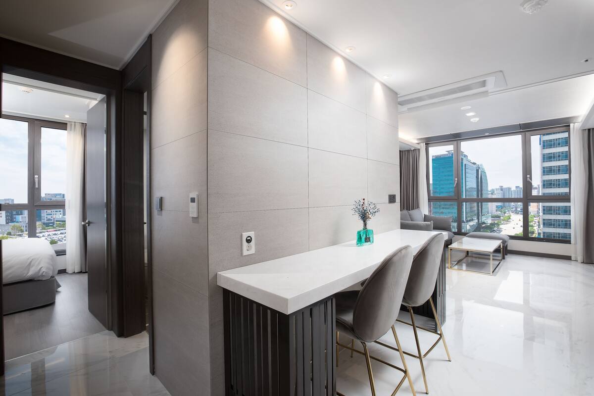 A modern hotel room with a large window, a dining area, and a bed.