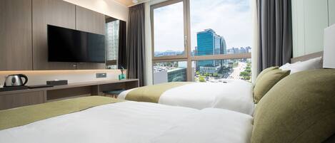A hotel room with a large bed, a flat-screen TV mounted on the wall, a desk with a kettle, and a view of a cityscape through large windows.