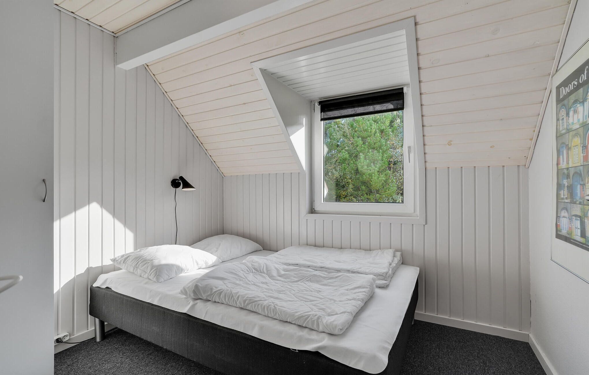 Et soveværelse med skråvæg, et vindue med udsigt over grøn vegetation, en seng med hvide sengetøj og en lampe monteret på væggen.