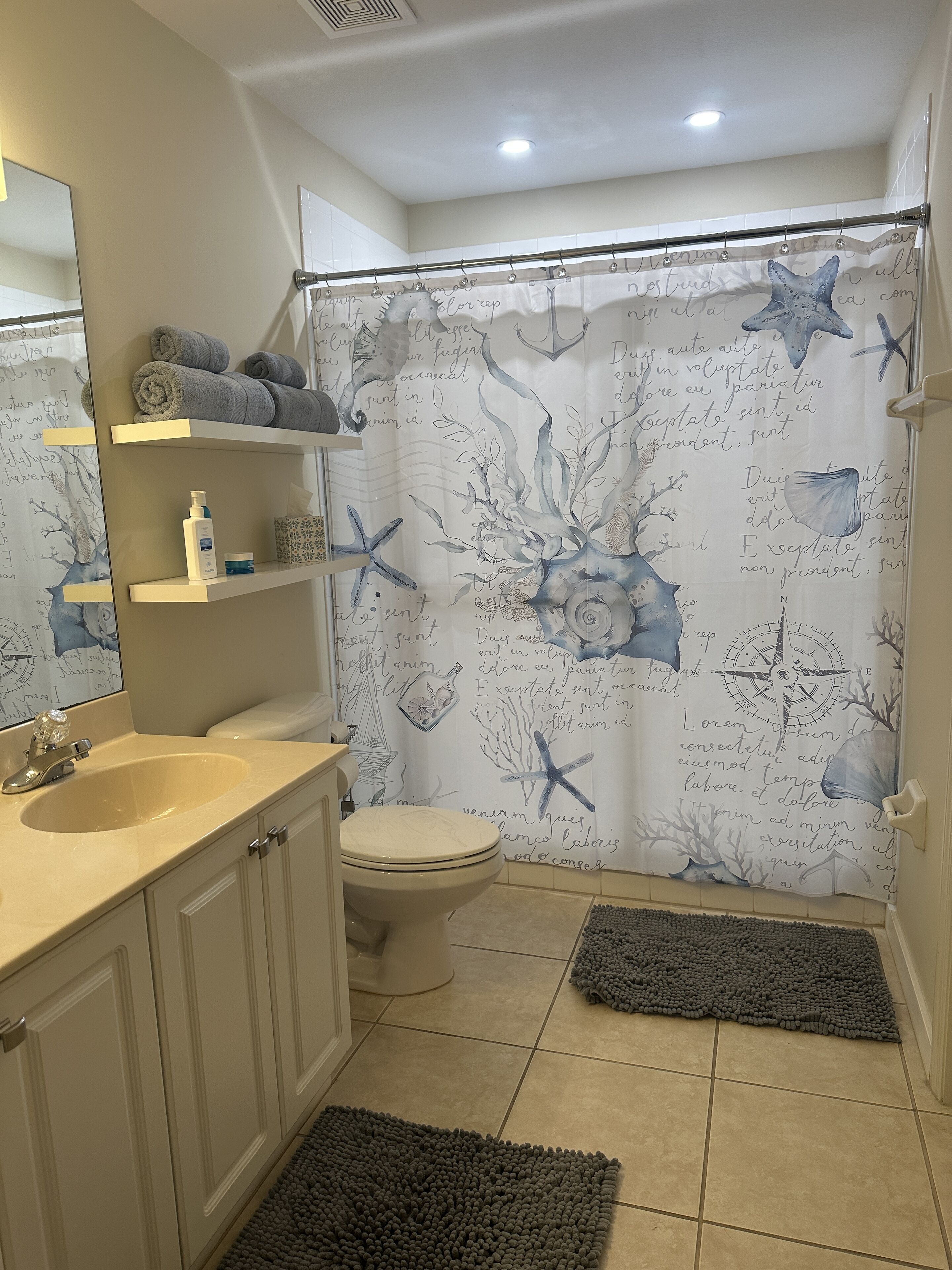 A bathroom with a beige sink, white toilet, and a shower curtain with sea-themed designs. There are shelves with towels and toiletries.