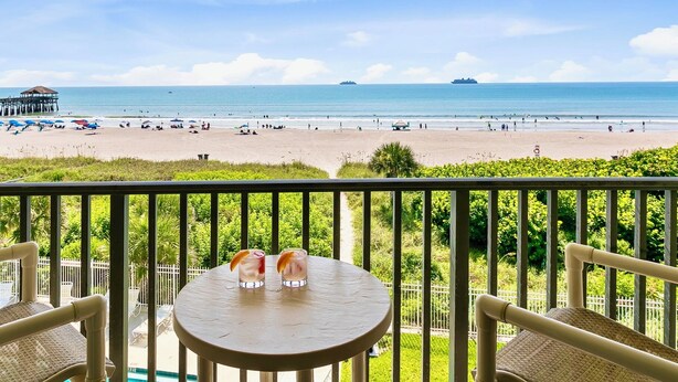 A balcony with a table and chairs overlooking a beach with umbrellas and people. - Oceanview Condo w/ Balcony – Steps to Cocoa Beach Pier! (Cocoa Beach)