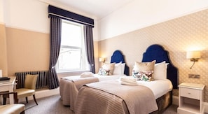 A hotel room with two beds, a desk, a chair, a radiator, and a window with curtains. - Queen s Hotel (Portsmouth)