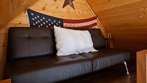 A cozy attic room with a wooden sloped ceiling, a black leather sofa, a white pillow, and a large American flag. - Treehouse Place at Deer Ridge Ole Mill (Snohomish)