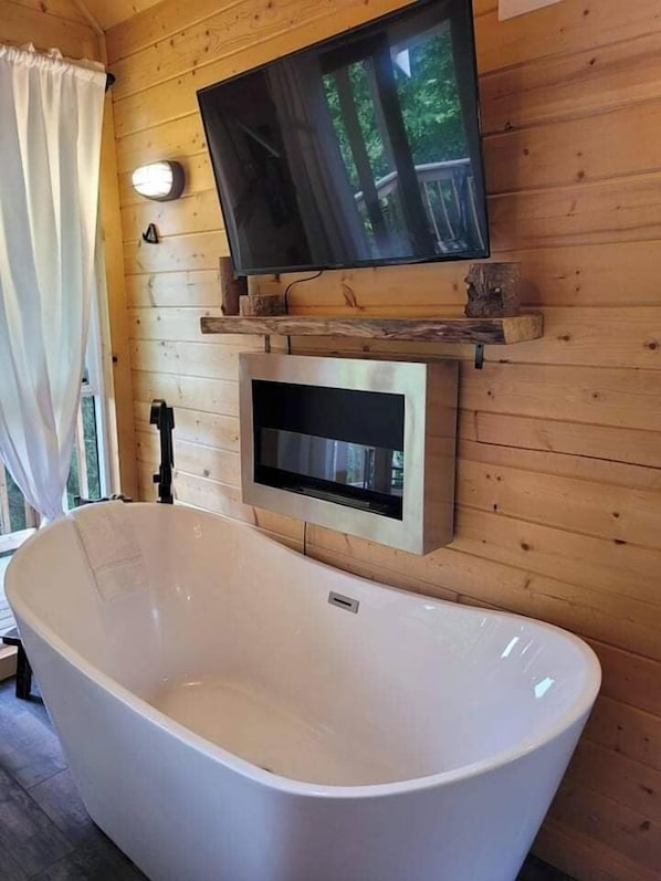 Interieur van een houten hutje met een vrijstaand bad, een bevestigde televisie en een raam met gordijnen.