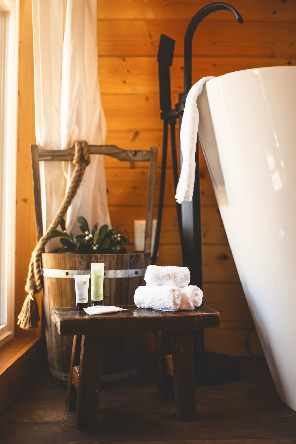 A wooden cabin interior with a bathtub, a chair with a rope, and a small table with towels and bottles.