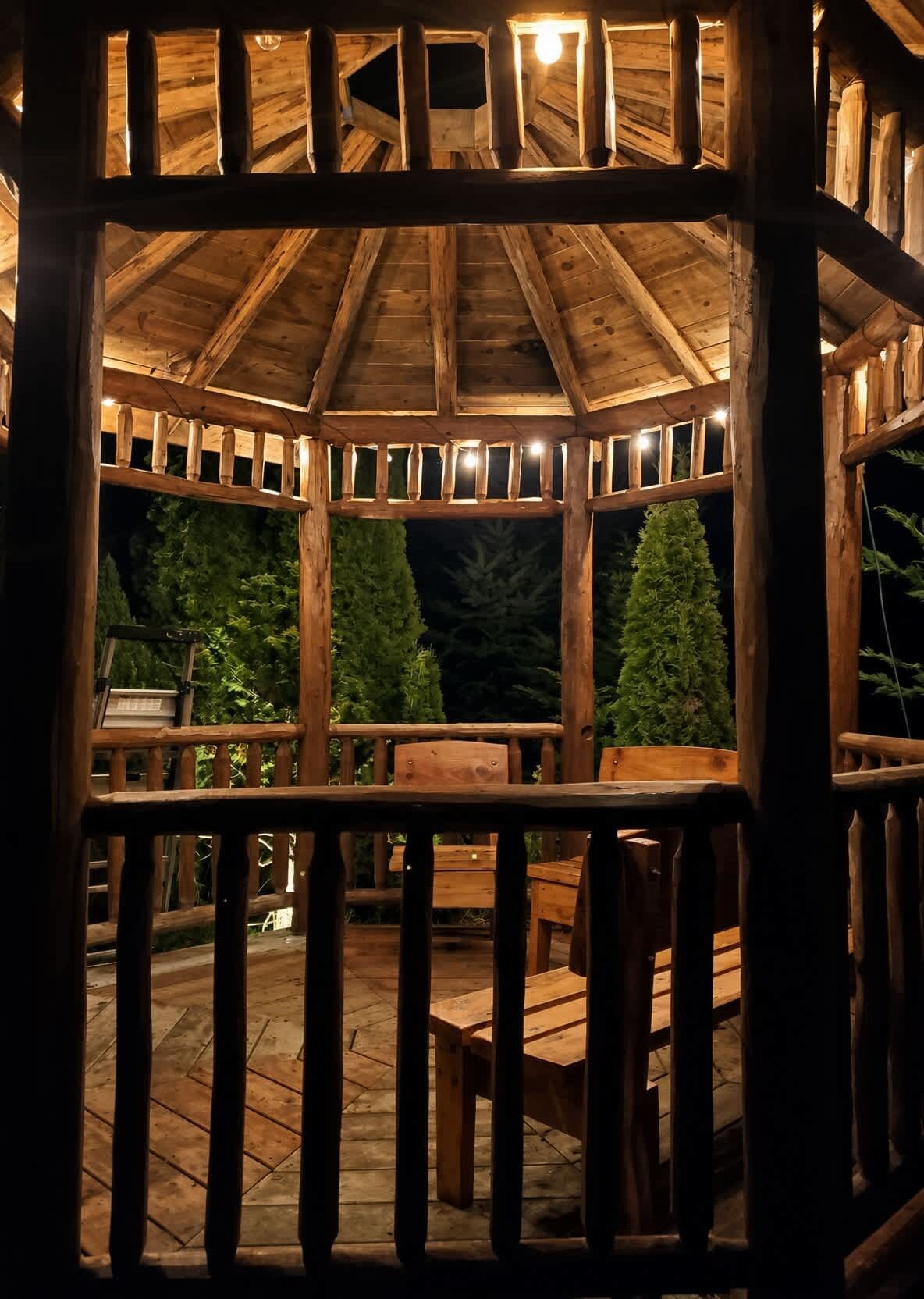A wooden gazebo with a roof, surrounded by a wooden deck and chairs.