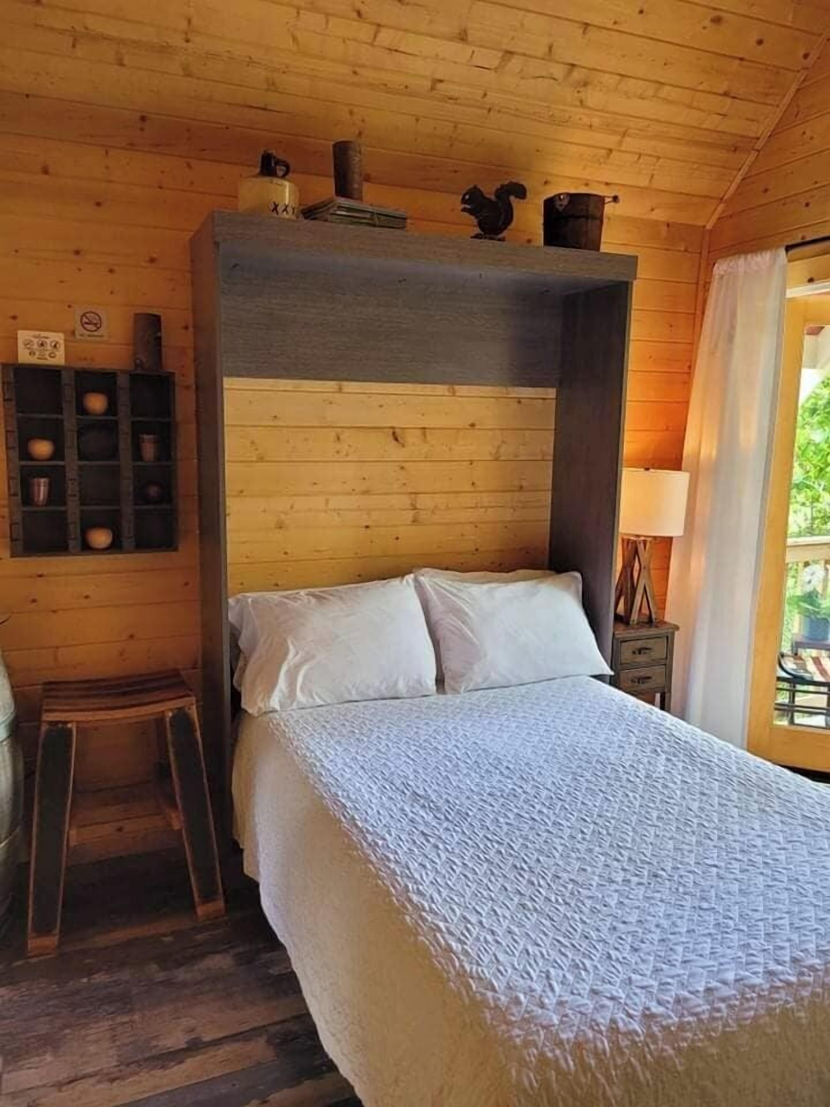A cozy wooden cabin room with a bed, a small stool, a shelf with items, and a window with a view.