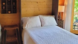 A cozy wooden cabin room with a bed, a small stool, a shelf with items, and a window with a view. - Treehouse Place at Deer Ridge Ole Mill (Snohomish)