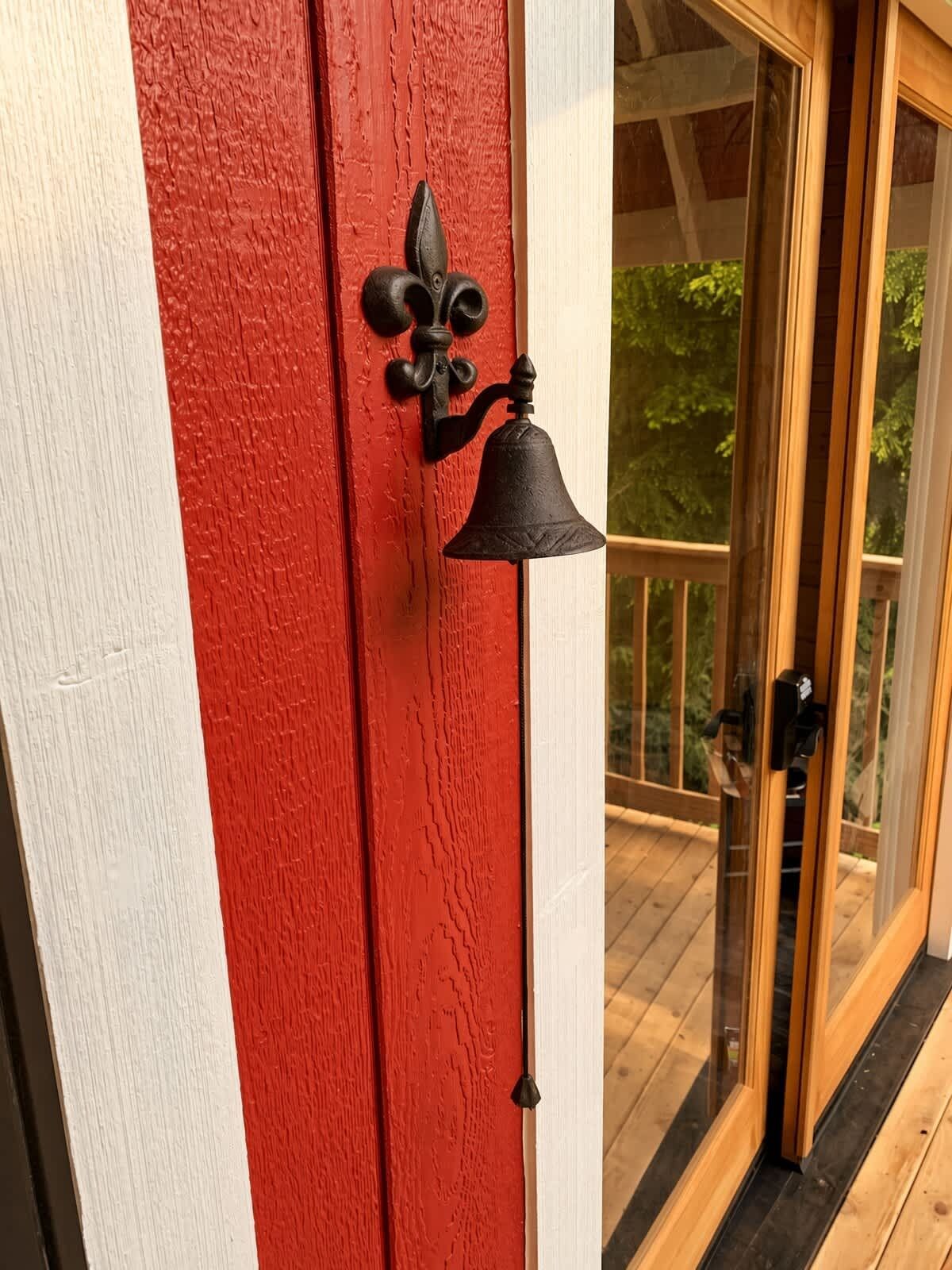 Une porte rouge avec une sonnette noire et un heurtoir, qui donne sur une terrasse en bois dotée de portes vitrées.
