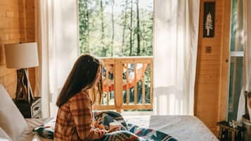 A cozy cabin room with a bed, a window with curtains, a wooden ceiling, and a small table with a lamp.