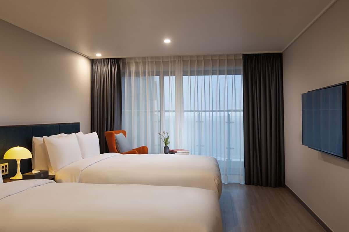 A hotel room with two beds, a nightstand with a lamp, a chair, a small table with a vase, and a large window with curtains.