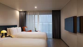 Habitación de hotel con dos camas, una mesita de noche con lámpara, una silla, una pequeña mesa con un jarrón y un ventanal con cortinas.