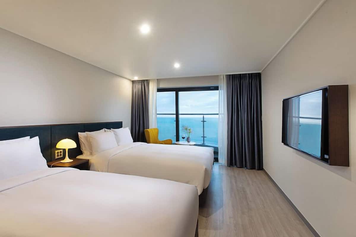 A hotel room with two beds, a nightstand with a lamp, a chair, a TV, and a large window with a view of the sea.