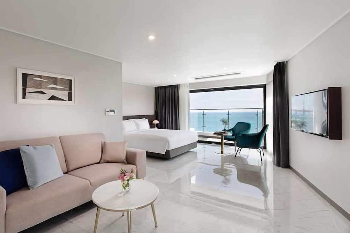 A modern hotel room with a bed, a sofa, a small table, and a TV mounted on the wall.