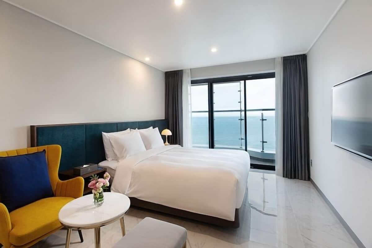 A hotel room with a bed, a yellow armchair, a small table with a vase, a TV, and a large window with a view of the sea.
