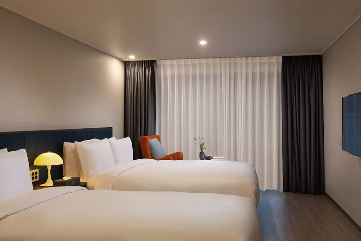 A hotel room with two beds, a nightstand with a lamp, a chair, a small table with a vase, and a TV mounted on the wall.