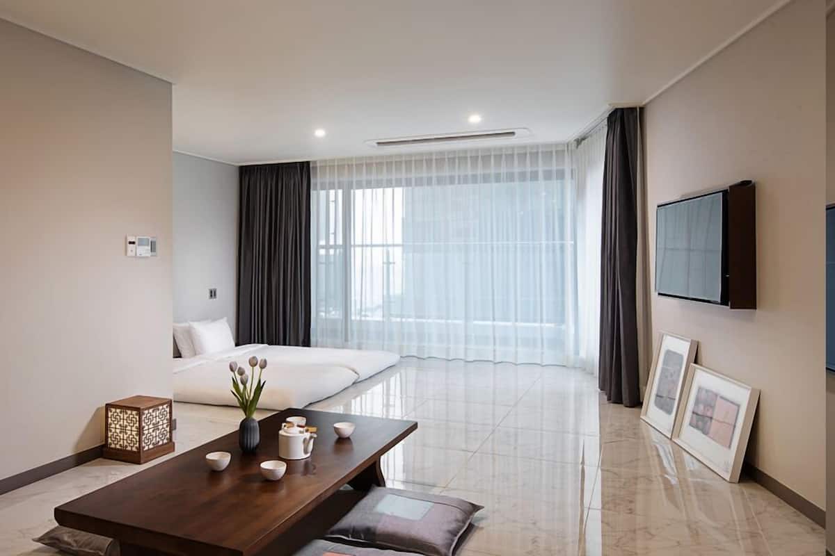 A modern hotel room with a bed, a dining table with tea set, a TV, and a large window with curtains.