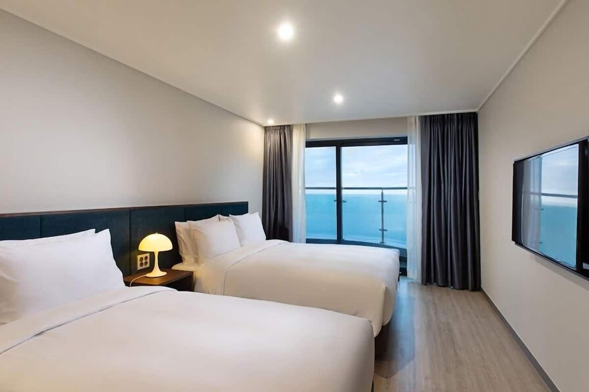 A hotel room with two beds, a nightstand with a lamp, a large window with curtains, and a TV mounted on the wall.