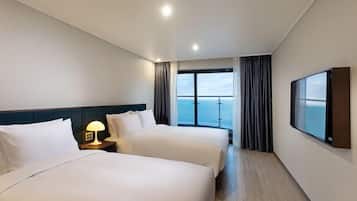 A hotel room with two beds, a nightstand with a lamp, a large window with curtains, and a TV mounted on the wall.