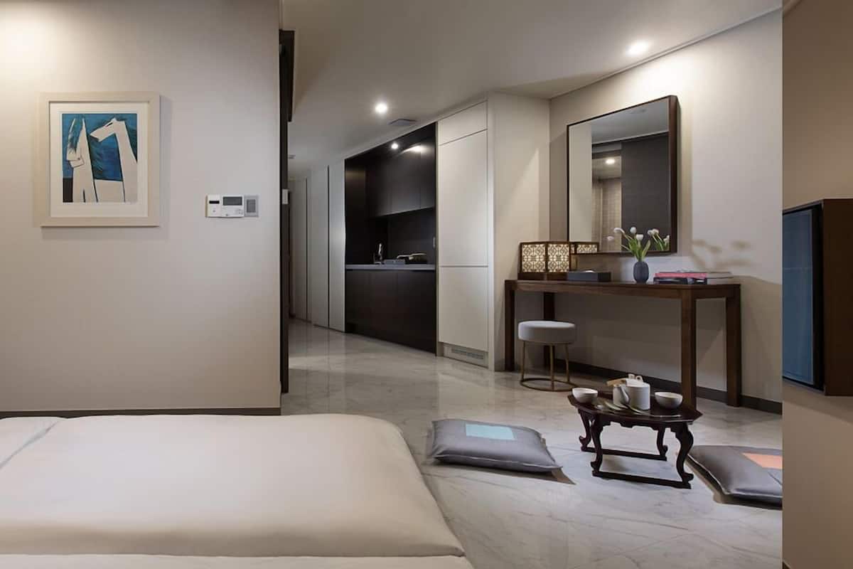 A modern hotel room with a bed, a desk, a chair, a small table with cups, and a painting on the wall.