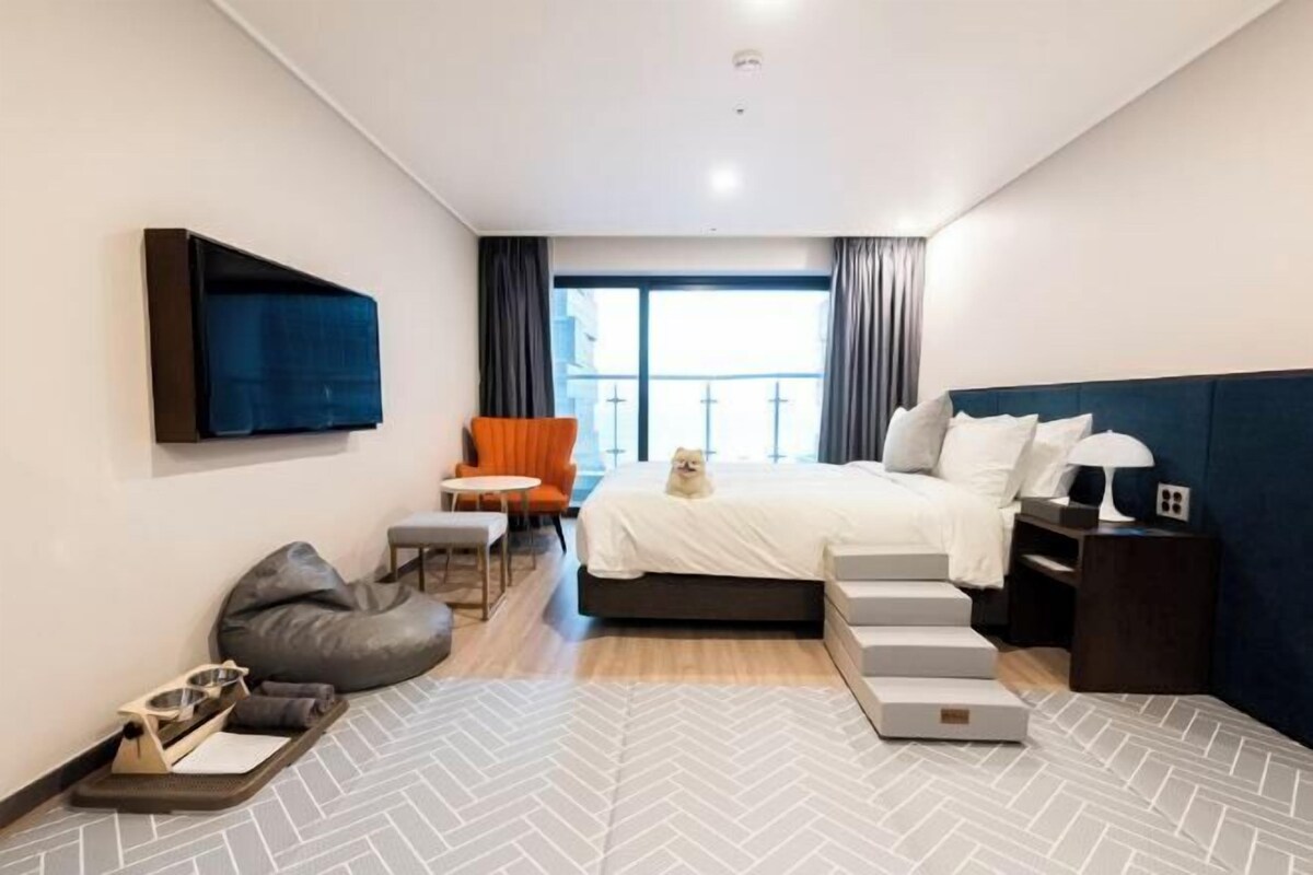 A modern hotel room with a bed, a TV mounted on the wall, a seating area with a chair and a small table, and a view of the outside through the window.