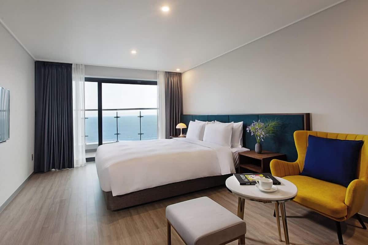 A hotel room with a bed, a yellow armchair, a small table, a nightstand, and a view of the ocean.