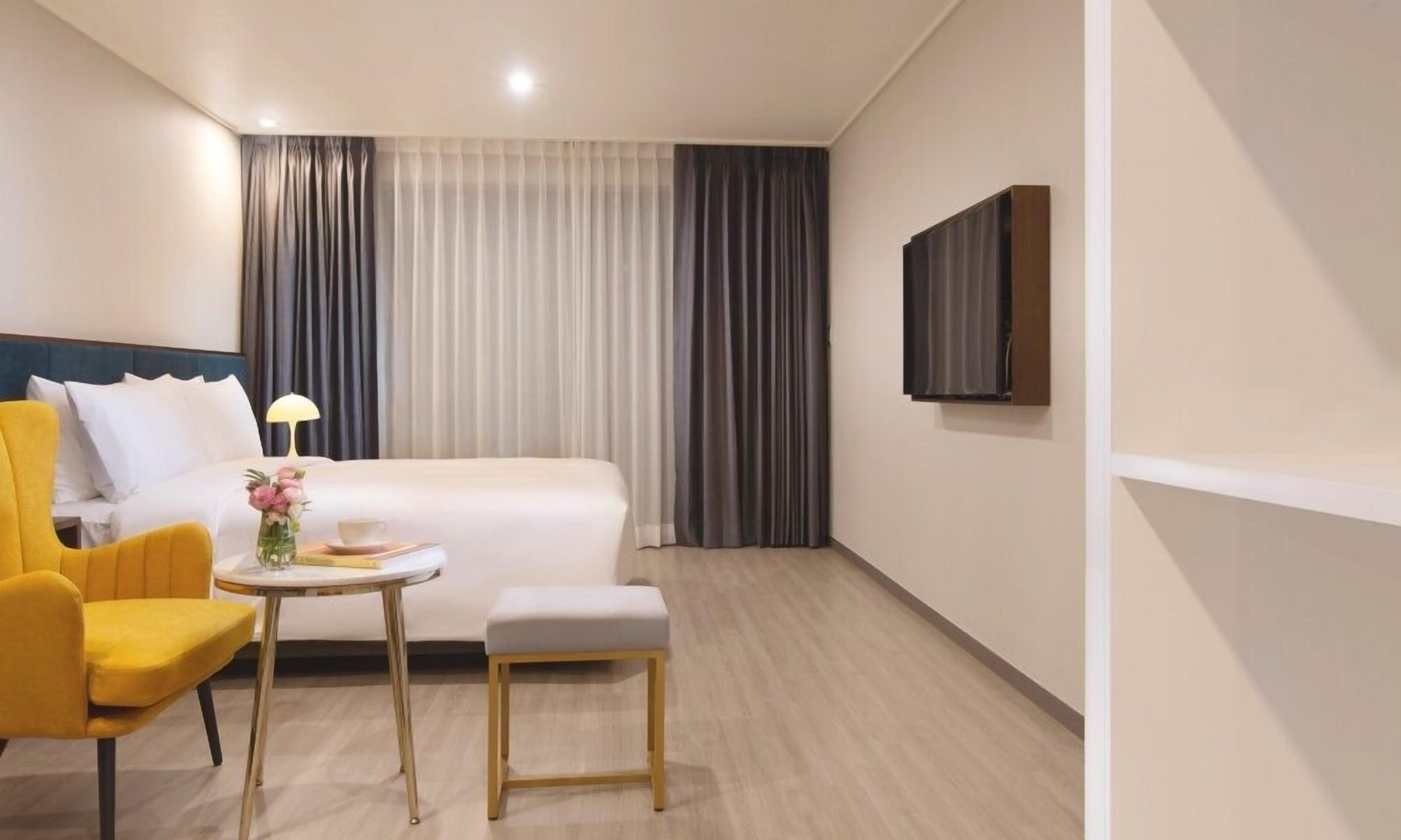 A hotel room with a bed, a small table with a lamp, a chair, and a TV mounted on the wall.