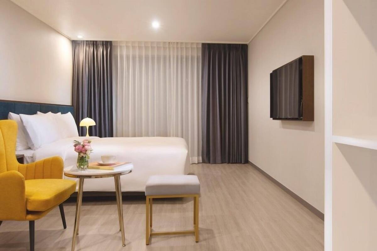 A hotel room with a bed, a small table with a lamp, a chair, and a TV mounted on the wall.