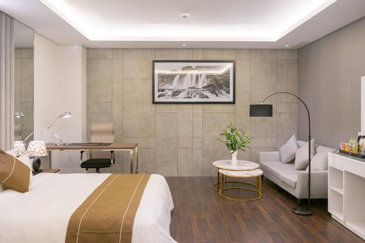 A modern hotel room with a large bed, a desk, a sofa, and a small table with a plant.