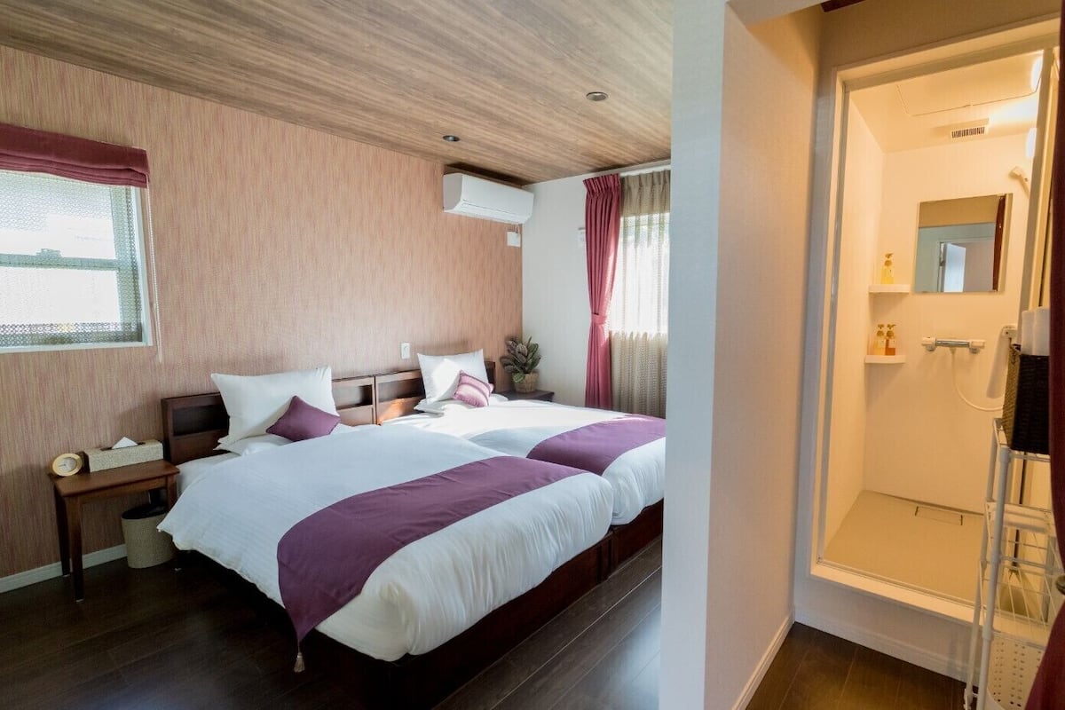 A hotel room with two beds, a bedside table, a nightstand, a window with curtains, and a bathroom with a shower.