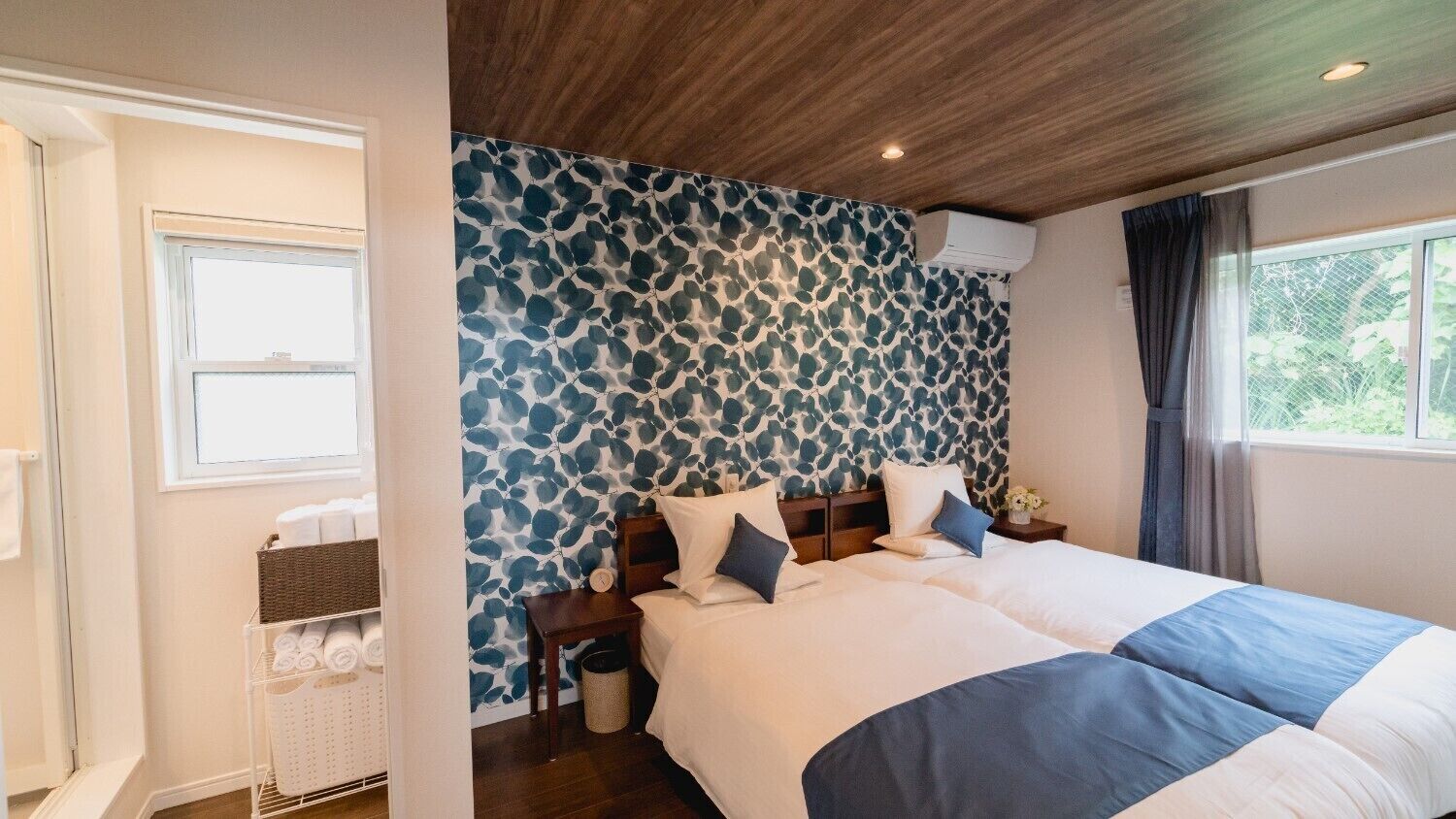 Een hotelkamer met twee bedden, een houten plafond, een behang met patroon, een raam en een nachtkastje met toiletartikelen.