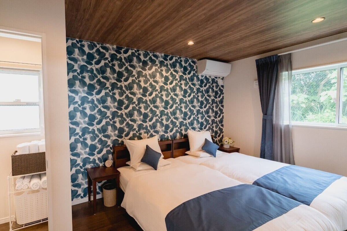 A hotel room with two beds, a wooden ceiling, a patterned wall, a window, and a nightstand with toiletries.