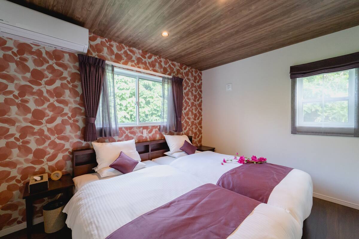 A bedroom with a bed, a nightstand, a wall-mounted air conditioner, and a window with curtains.