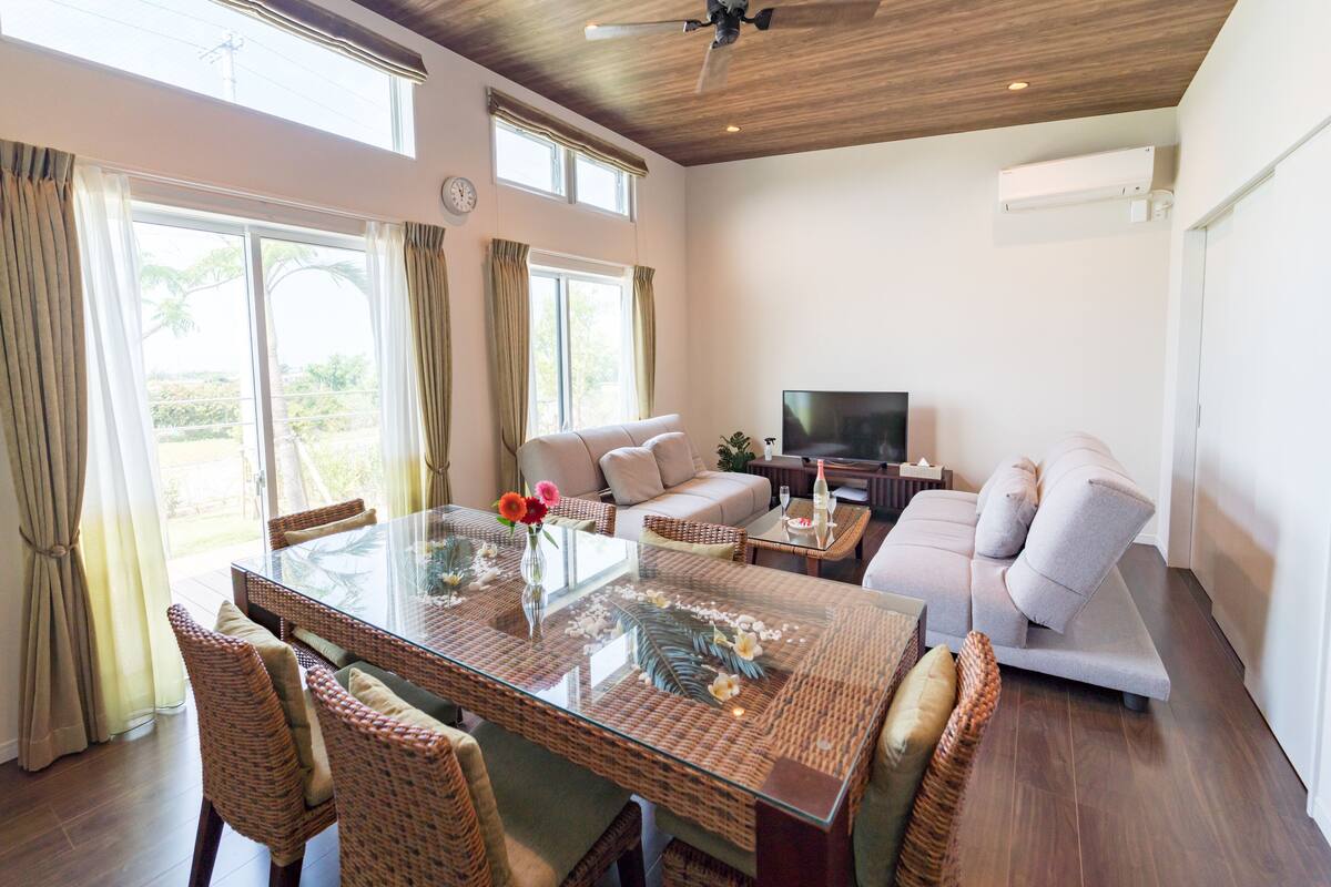 A spacious living room with a glass-top dining table, wicker chairs, a sofa, and a television.