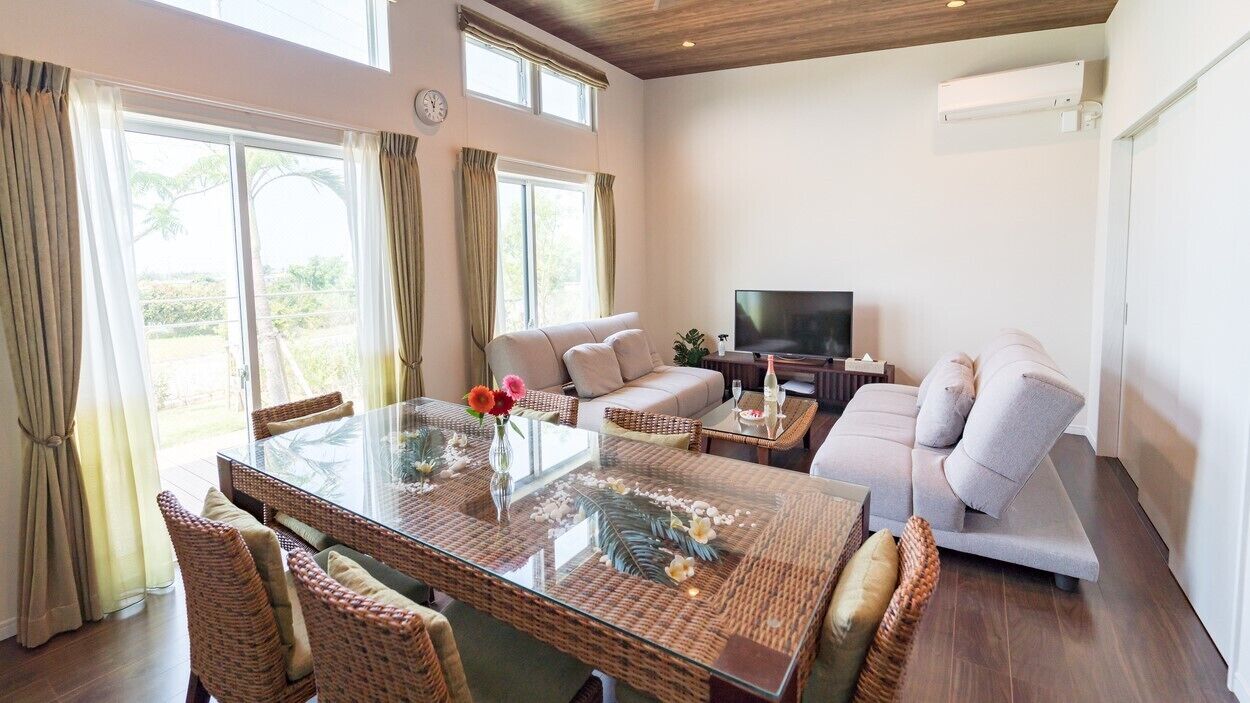 A spacious living room with a glass-top dining table, wicker furniture, a sofa, and a flat-screen TV.
