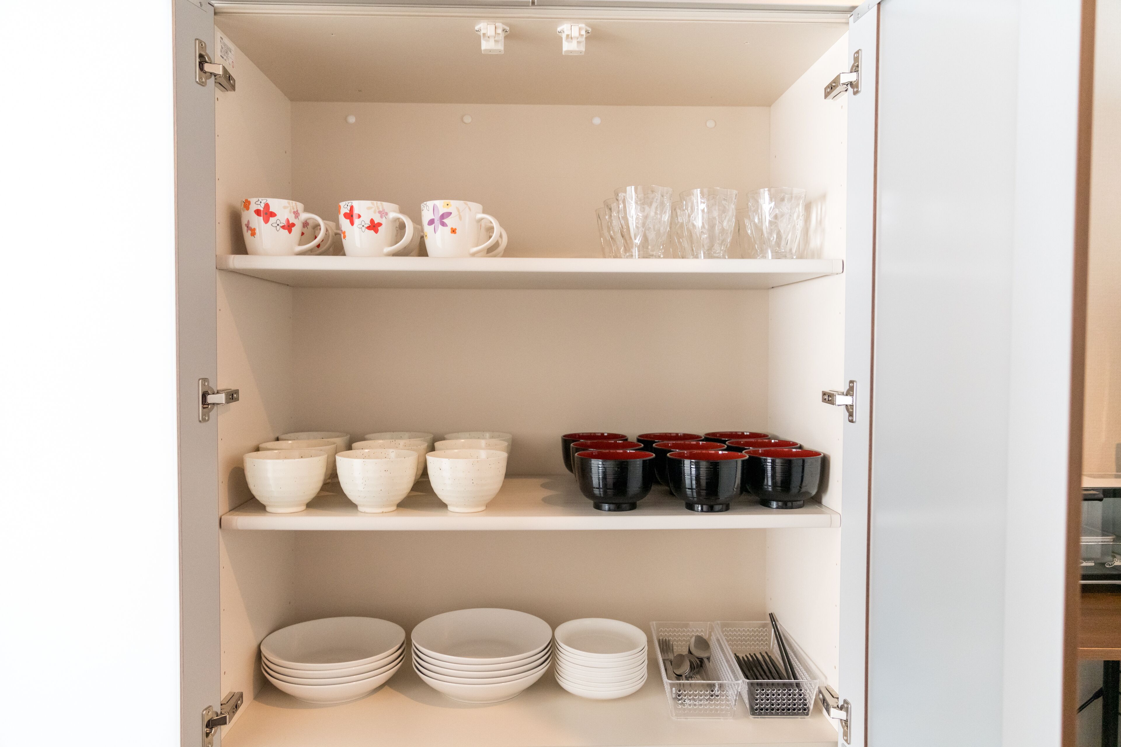 Un mueble de cocina con cuencos blancos y rojos, tazas blancas y rojas, y vasos transparentes.
