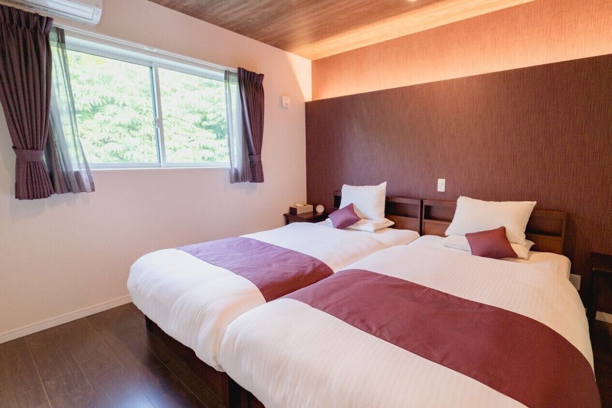A double bed with a headboard, a bedside table with a vase of flowers, and a window with curtains.