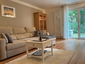 Ein Wohnzimmer mit einem beigefarbenen Sofa, einem weißen Couchtisch mit einer Vase, einem Holzschrank und Blick durch eine Glastür.